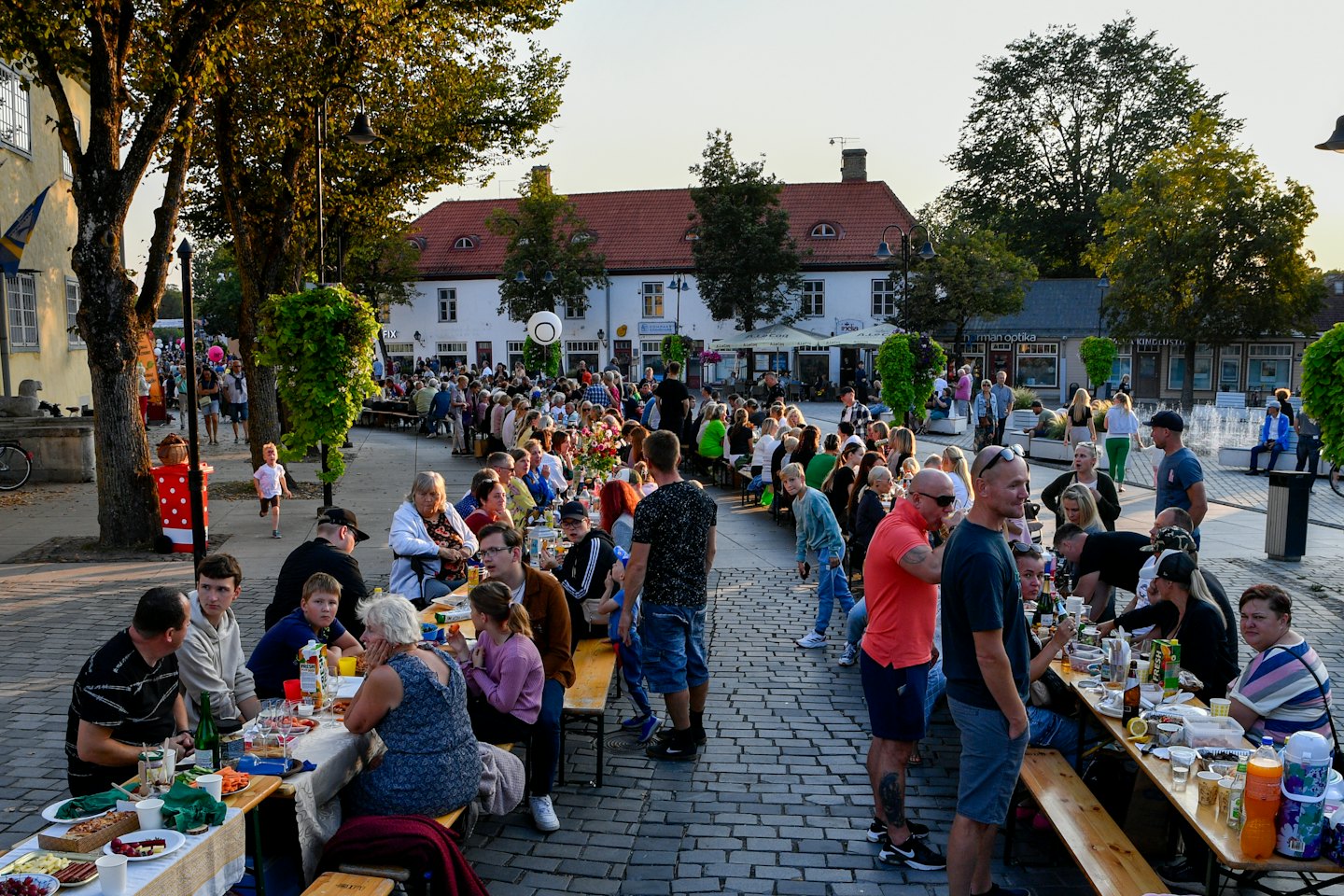 Persone che mangiano e bevono ai tavoli in una piazza cittadina al tramonto.